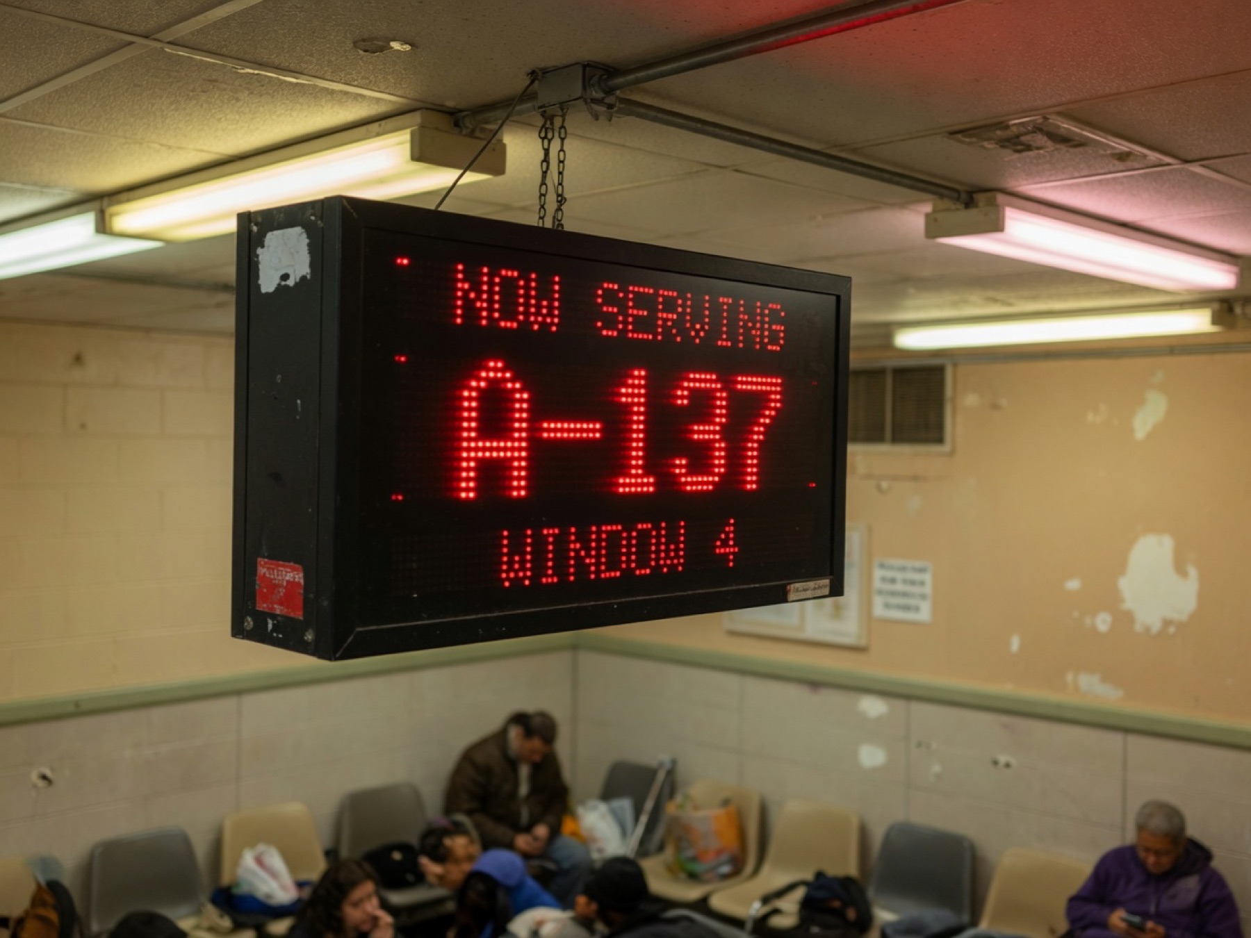 A hanging Now Serving LED display over a DMV waiting room.